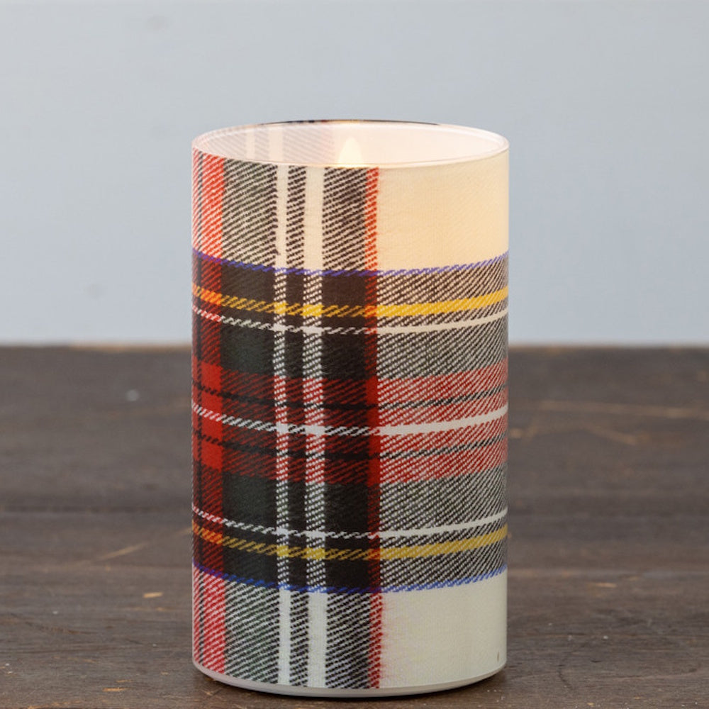 Lit white tartan plaid glass candle sitting on a wooden surface.