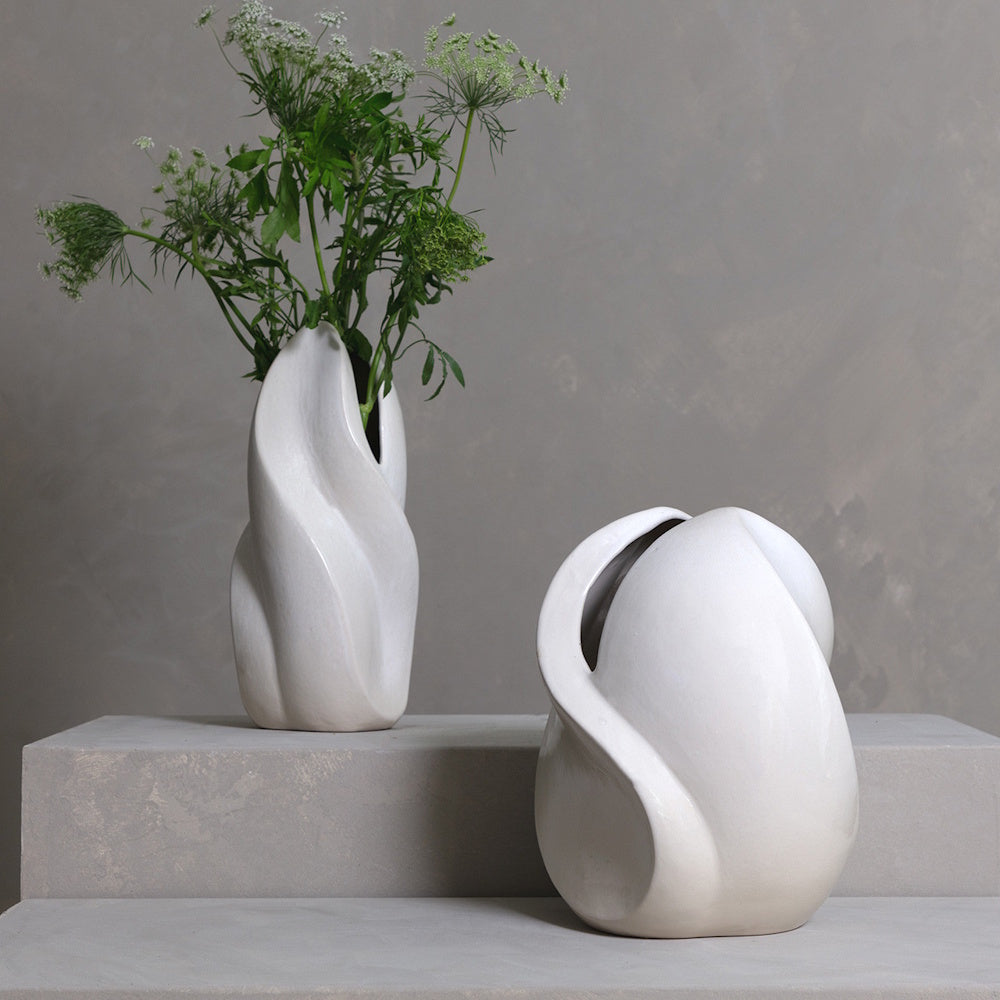 Two modern white ceramic twist vases on stone steps, one holding green botanical sprigs against a gray wall.