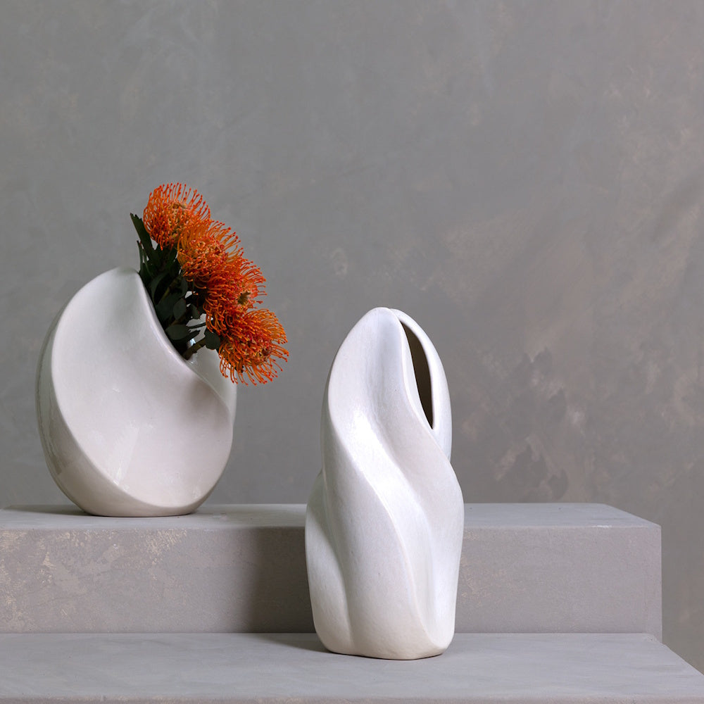 Two modern white sculptural ceramic vases on a stone ledge, one holding orange pincushion protea flowers.
