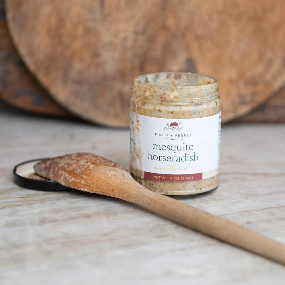An open jar of Finch + Fennel Mesquite Horseradish Mustard on a marble counter with a wooden spoon.