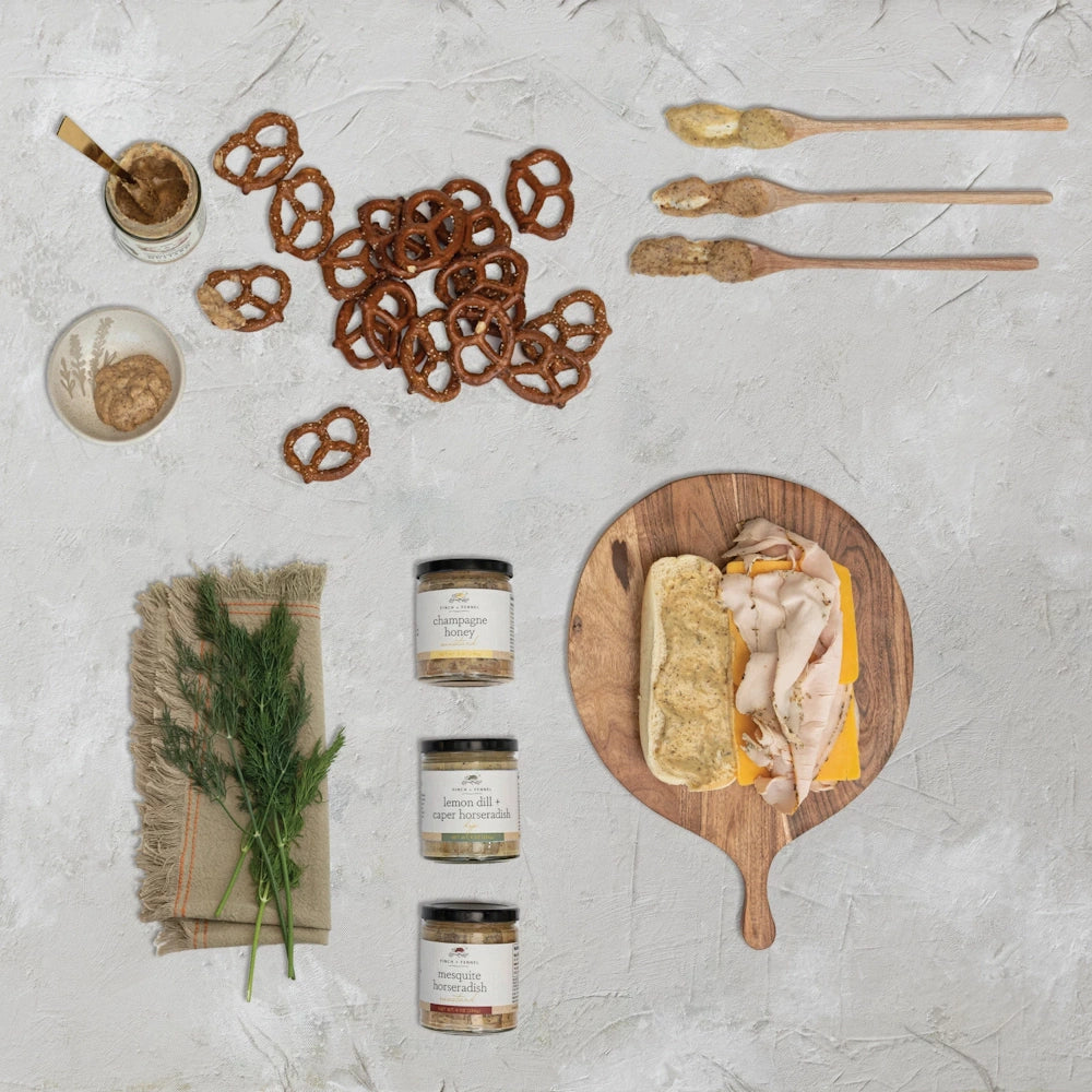 Gourmet mustard jars, pretzels, and a turkey sandwich on a wooden board with fresh dill on a grey textured background.