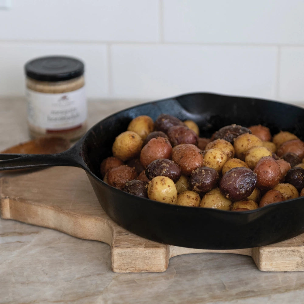 Roasted baby potatoes in a cast iron skillet next to a jar of mesquite horseradish mustard on a wooden board.