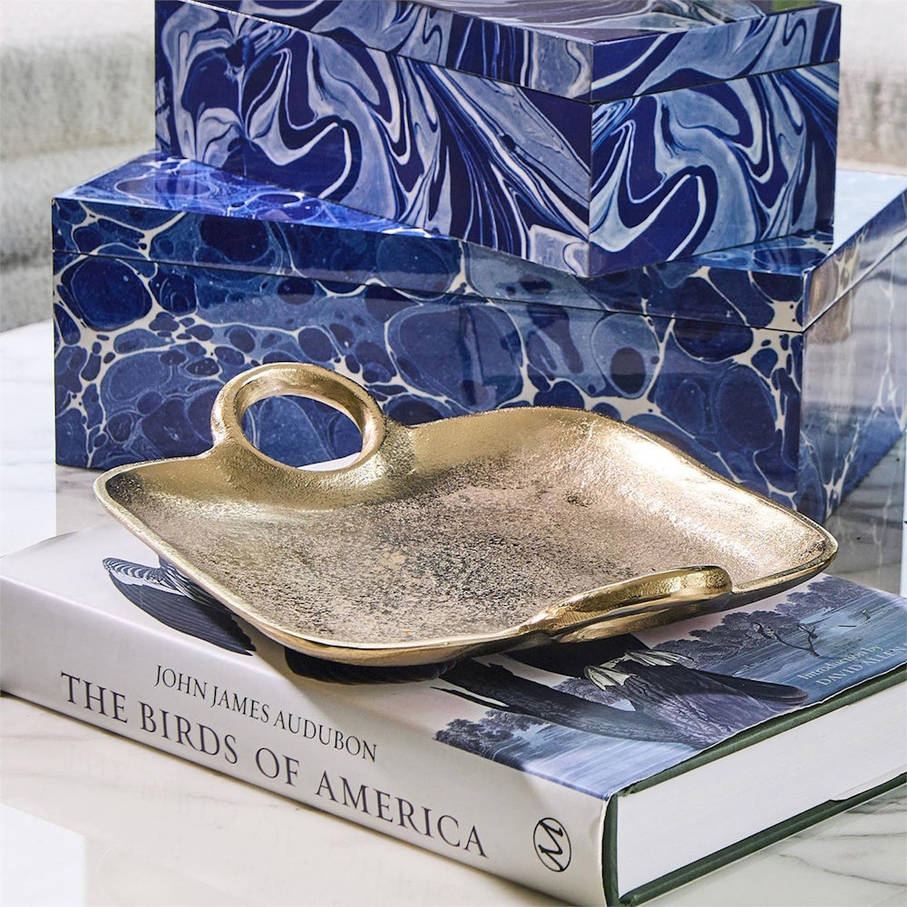 Textured gold decorative tray with handles resting on a book next to marbled blue boxes in a styled setting.