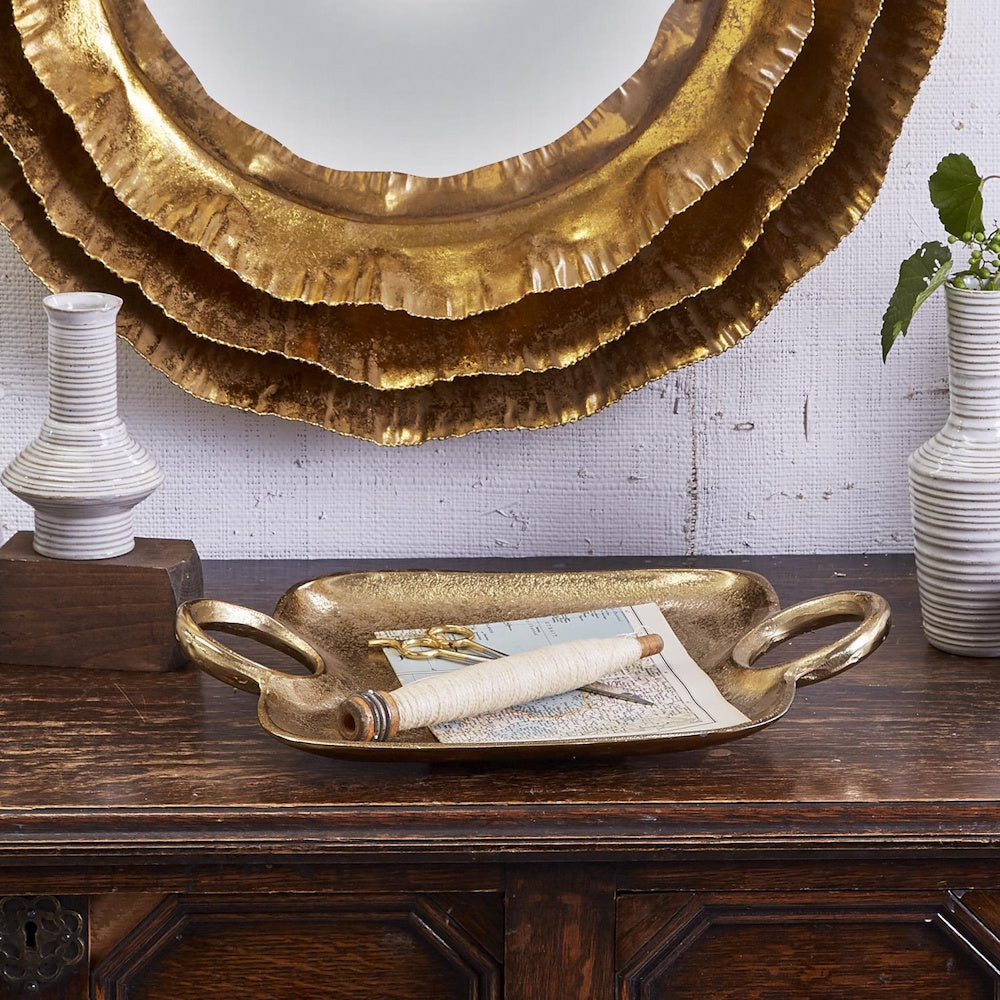 Textured gold decorative tray with handles styled on a wooden console below a layered gold mirror.