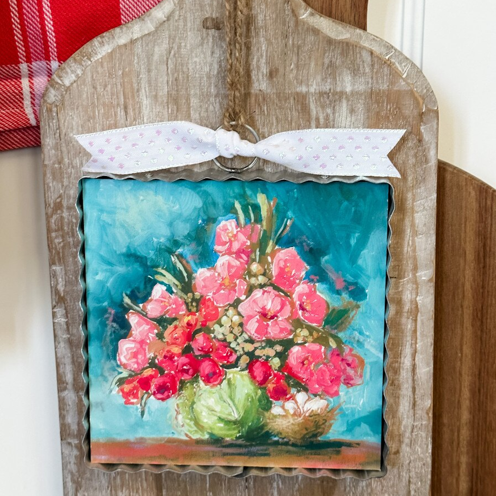 Floral painting of pink flowers in a cabbage vase on a corrugated metal sign with a white bow and twine hanger.