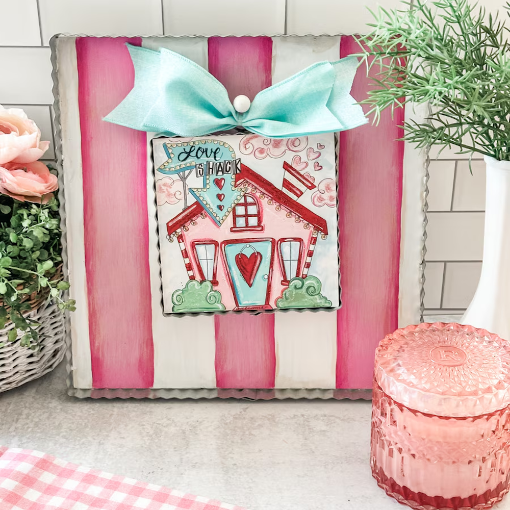 "Love Shack" whimsical house art on pink striped metal frame with teal bow, styled with pink decor and greenery.