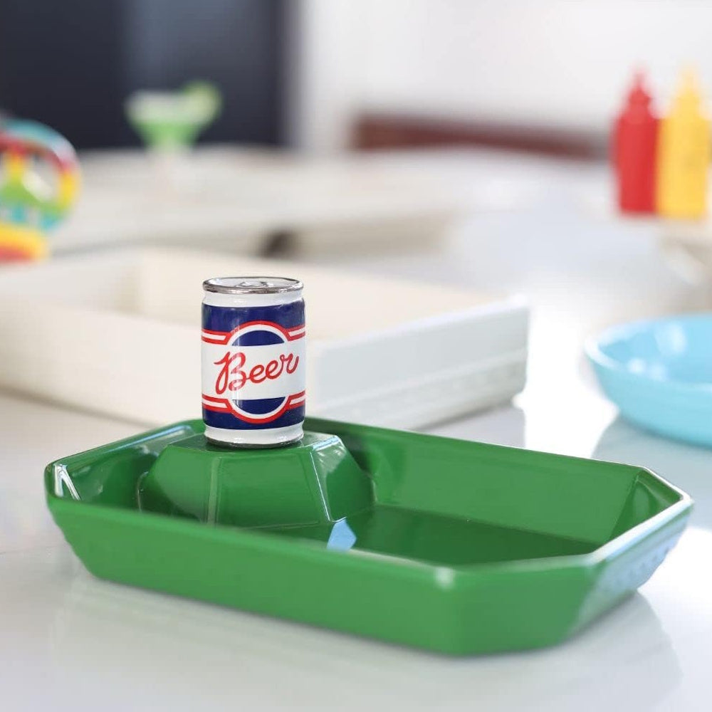 A miniature beer can sits on a raised platform inside a green rectangular dish on a tabletop.