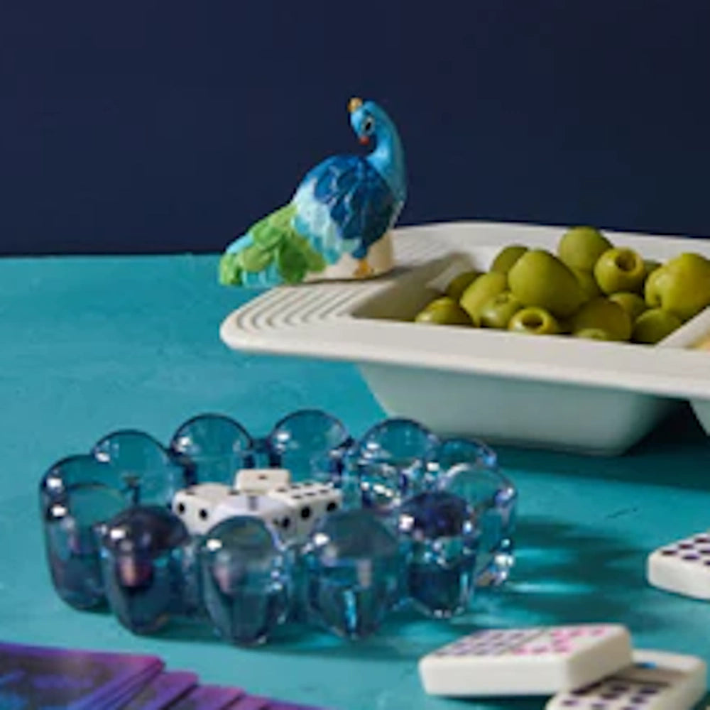 A fun peacock figurine perches on a snack bowl, with glass beads and dominoes arranged for a game on a blue table.
