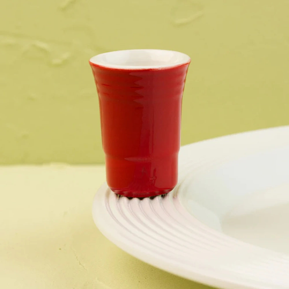 A miniature red ceramic party cup charm attached to the rim of a white textured plate.