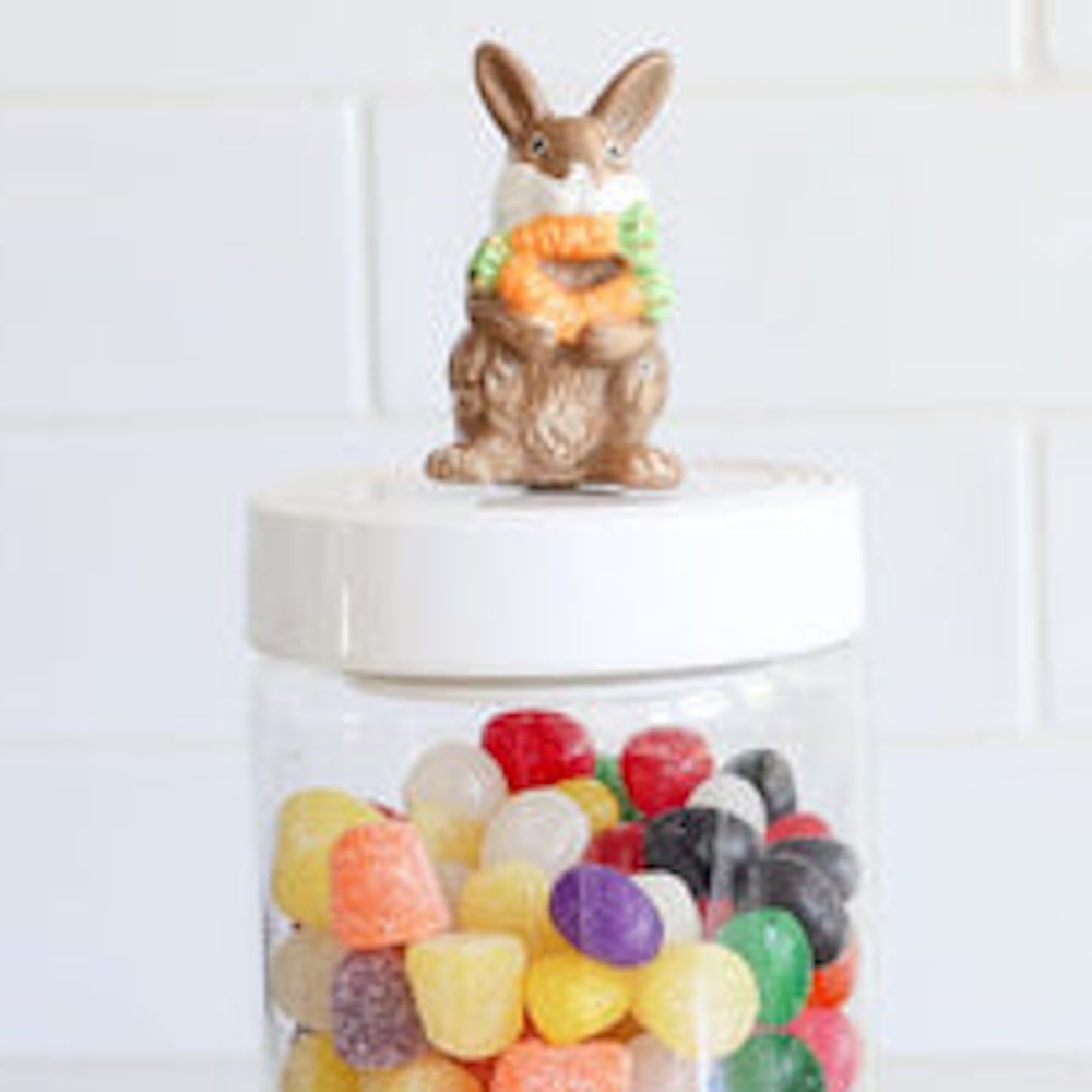 A small bunny figurine holding a small carrot sits on top of a jar filled with various colorful jelly beans.