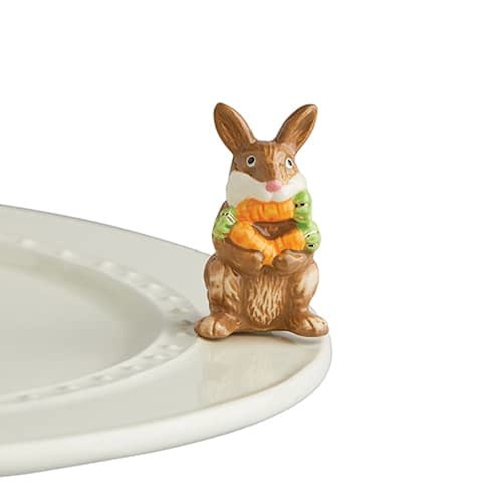 Ceramic brown bunny figurine holding orange carrots, perched on the edge of a white decorative serving platter.