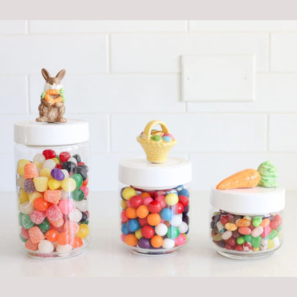 Three Easter candy jars on a counter, topped with a bunny, egg basket, and carrot figurines.