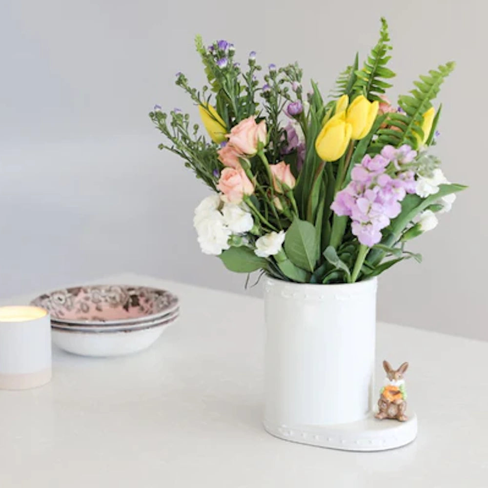 Spring floral arrangement in a white ceramic bunny vase on a white table with a lit candle and patterned bowls.