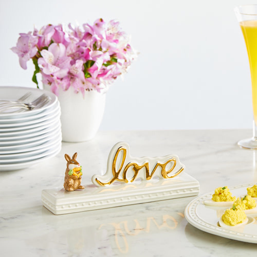 Ceramic love sign with a gold bunny figurine on a table with flowers, plates, and deviled eggs.