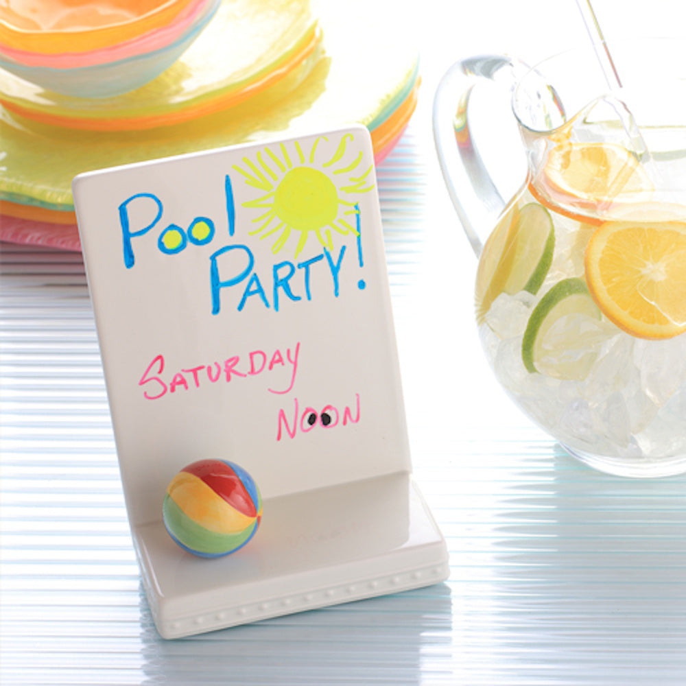 A ceramic white sign reading "Pool Party! Saturday Noon" next to a pitcher of citrus-infused ice water.