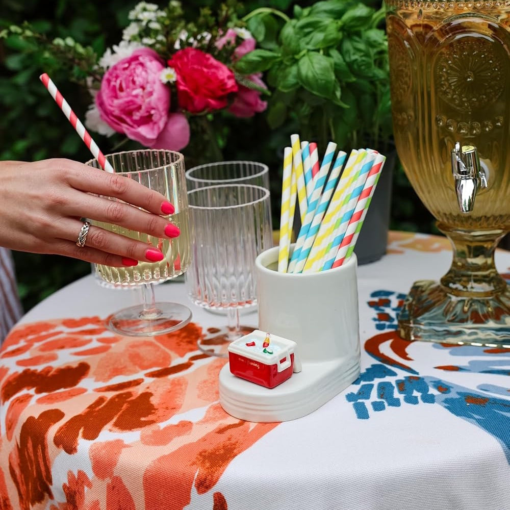 Hand reaching for a ribbed glass on a table with a white straw holder featuring a tiny red cooler and colorful straws.