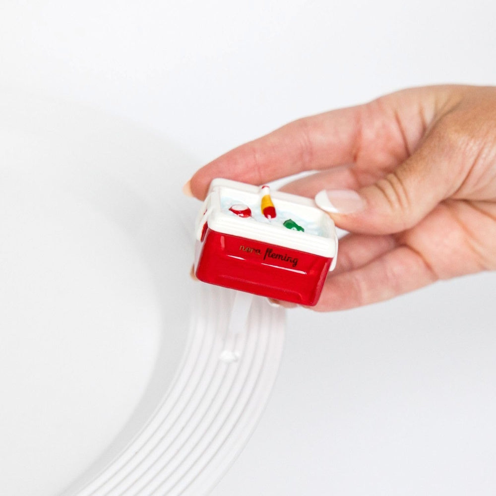 A hand placing a Nora Fleming "Ice Cold" mini red cooler charm into a white ceramic serving platter against a white background.