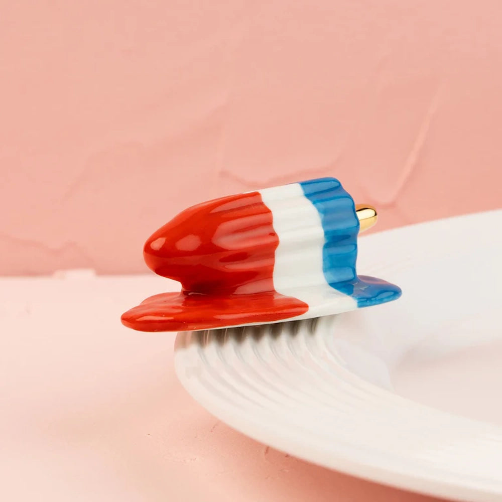 A red, white, and blue rocket popsicle ceramic figurine resting on the edge of a white plate.