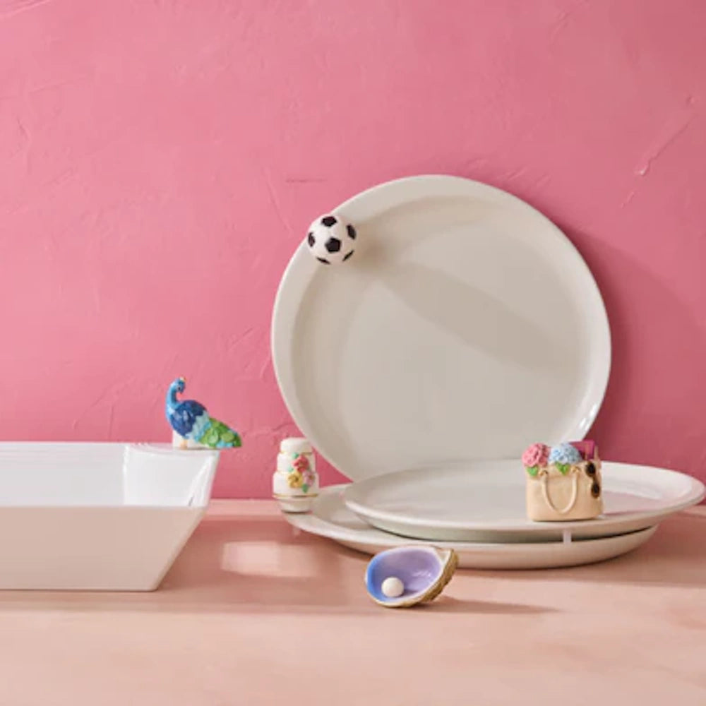 White ceramic plates and bowl with decorative soccer ball, peacock, and floral charms against a pink background.