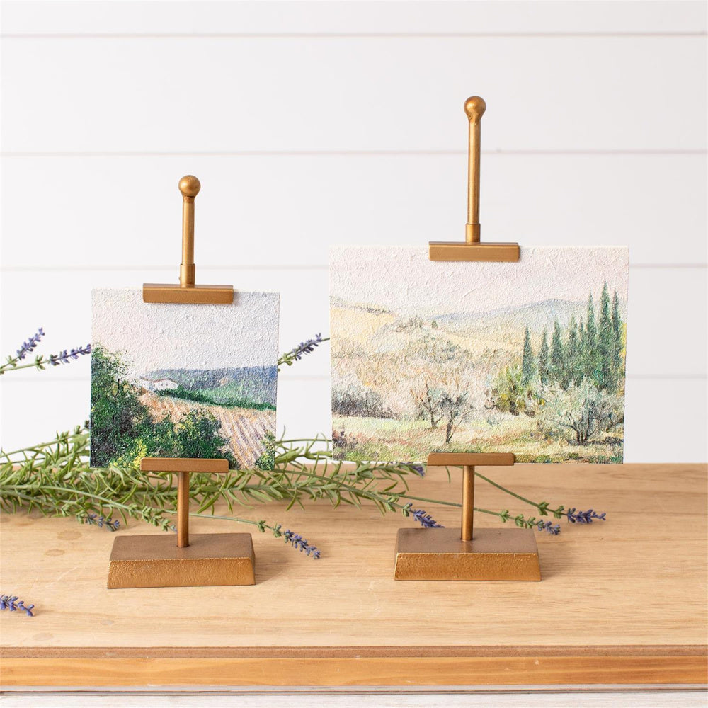 Miniature landscape oil paintings displayed on small gold easel stands with lavender sprigs on a wooden surface.