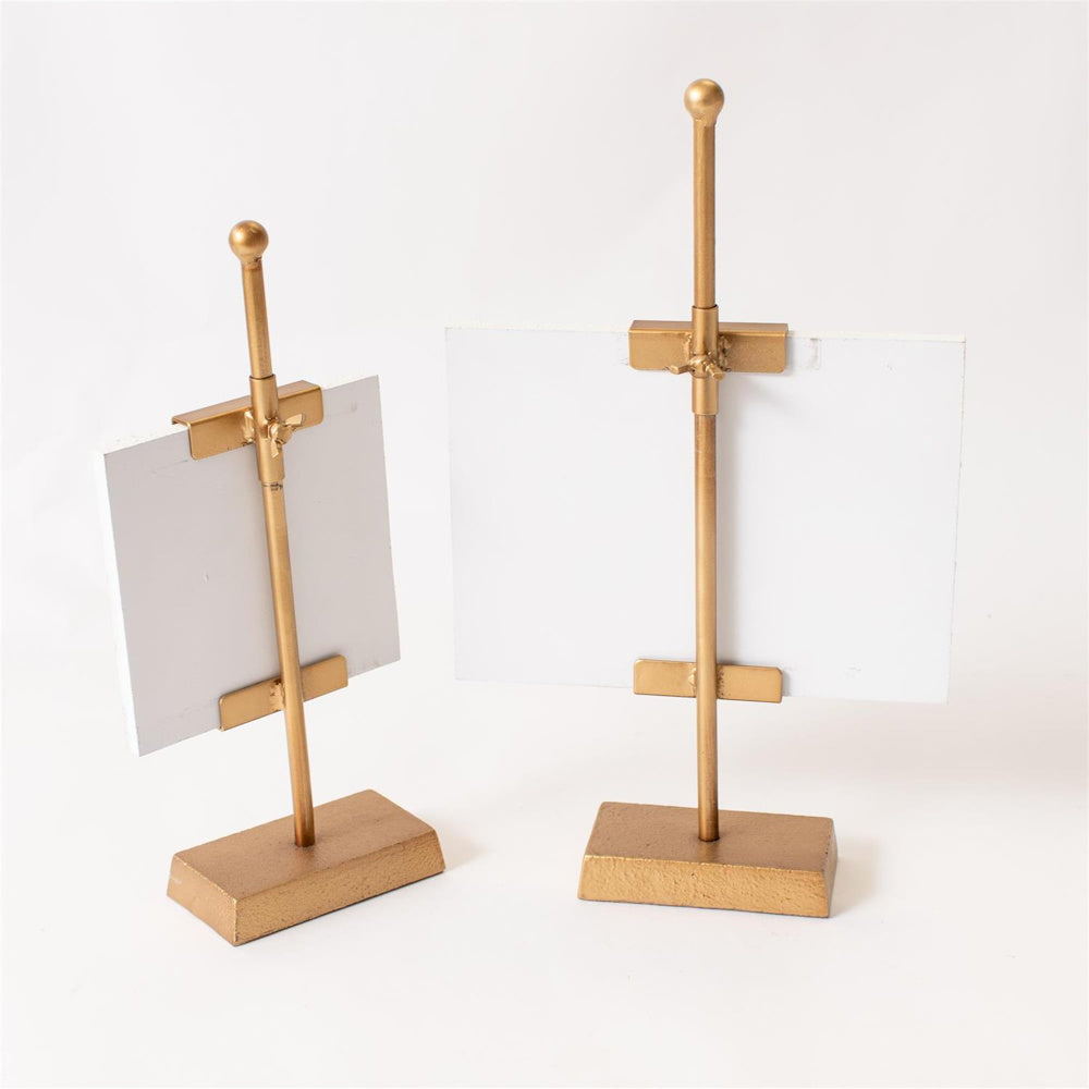 Two small gold metal easel stands holding white landscape art canvases on a white background.