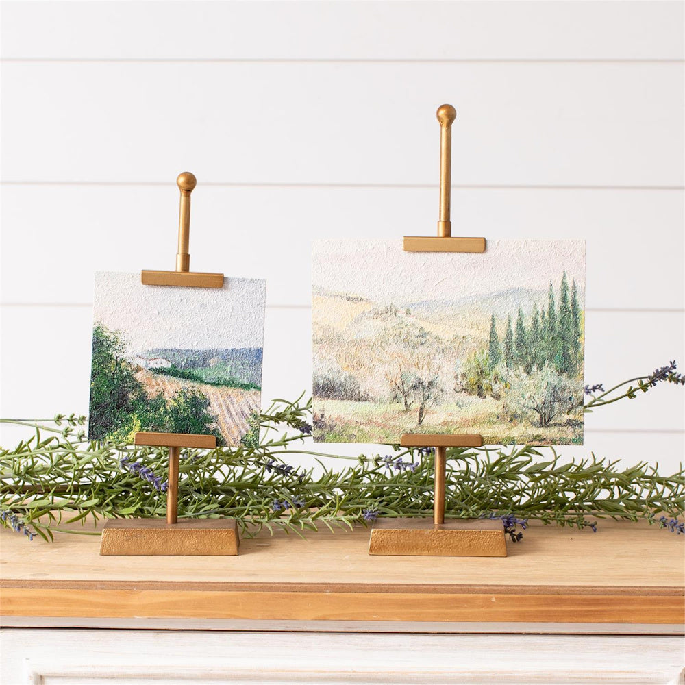 Two mini landscape paintings on gold easel stands displayed on a wooden surface with lavender sprigs and white wood backdrop.