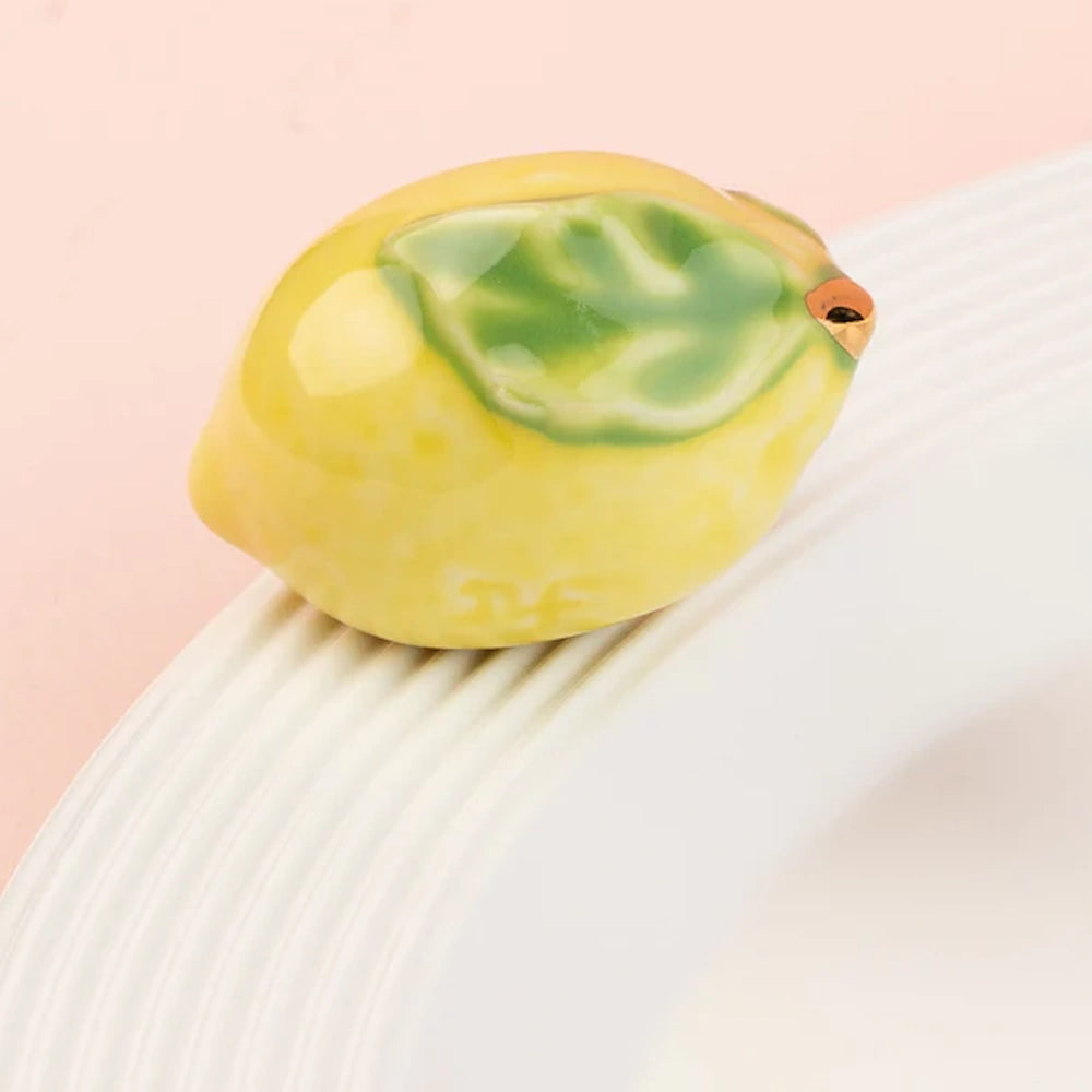 A small ceramic yellow lemon with a green leaf sitting on the rim of a white textured plate.