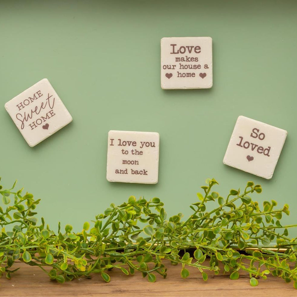 Set of four square cream magnets with love sayings and hearts, displayed on a green wall above artificial greenery.