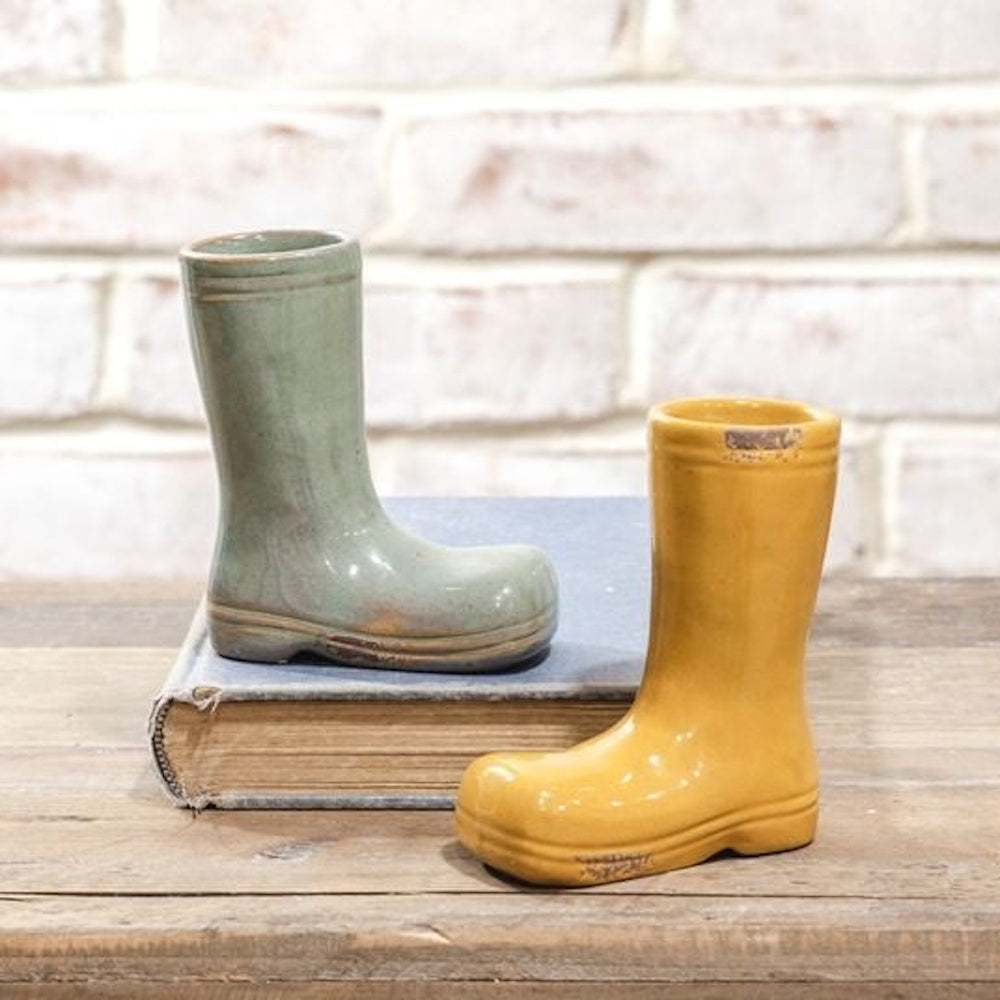 Two small ceramic rain boot planters, one green and one yellow, on a book against a brick background.
