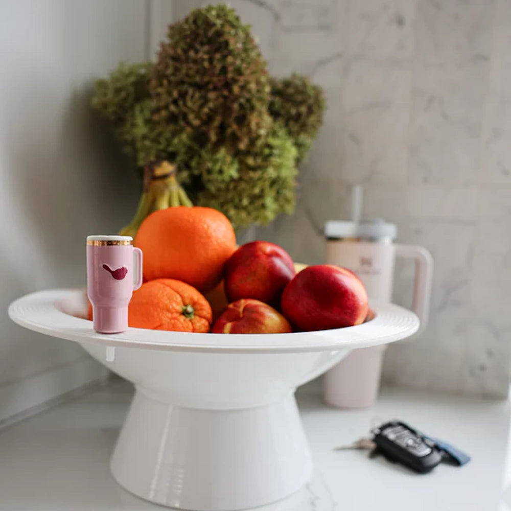 White pedestal fruit bowl filled with oranges and apples, featuring a miniature pink tumbler charm on the rim.