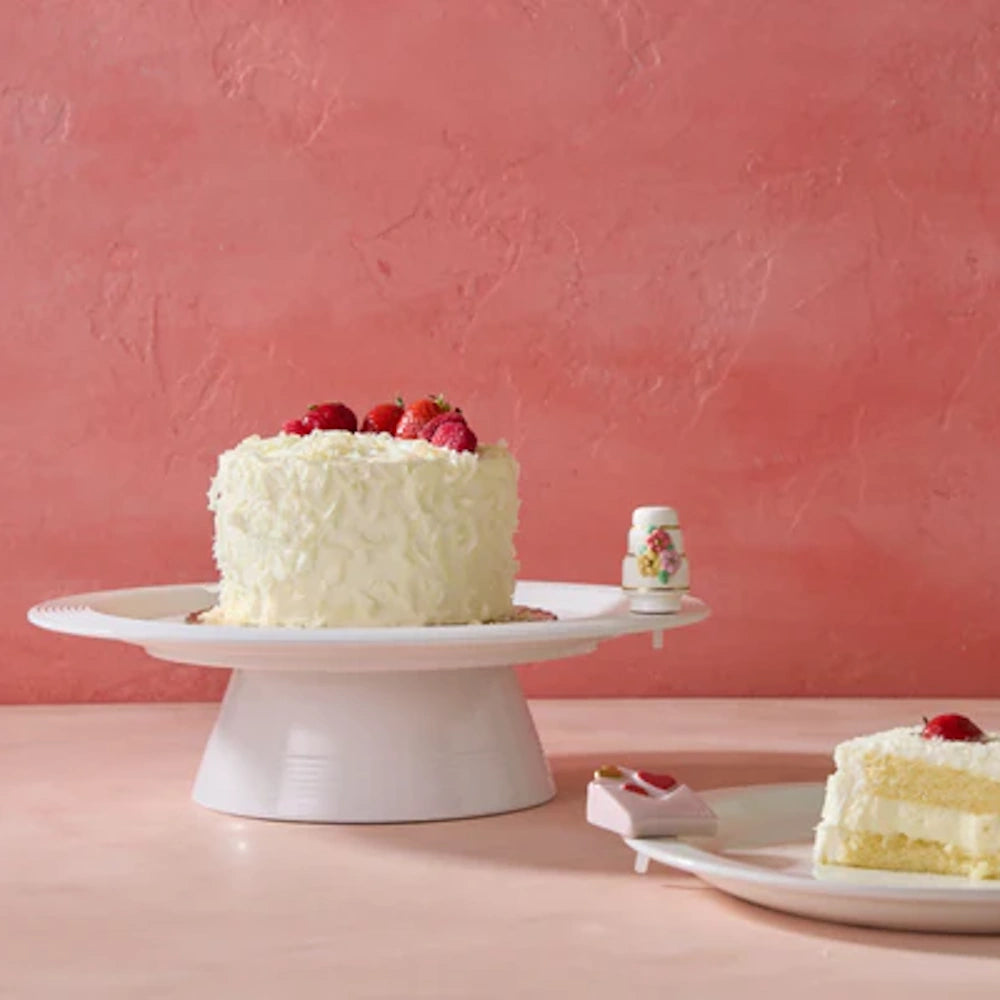 White cake with shredded coconut frosting and fresh strawberries on a white stand against a pink background.