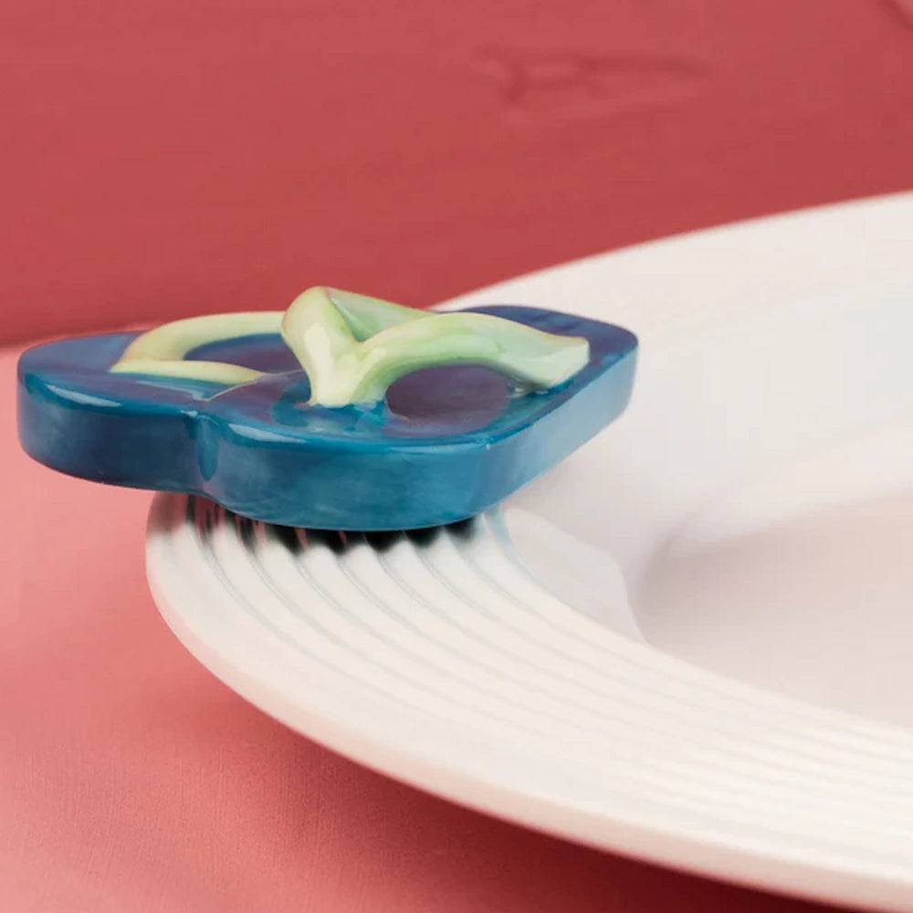 A tiny blue ceramic flip-flop charm attached to the rim of a white textured plate.