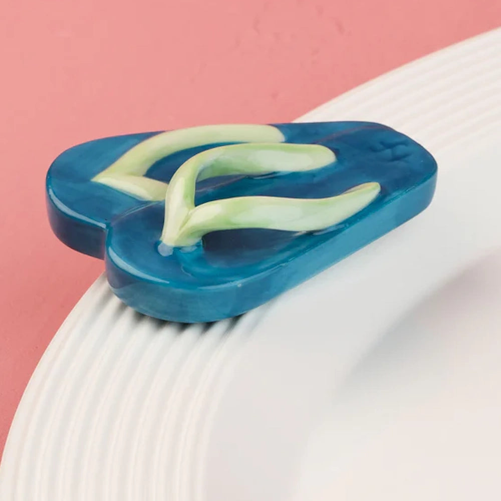 A small blue ceramic flip-flop charm with a light green strap resting on the edge of a white plate.