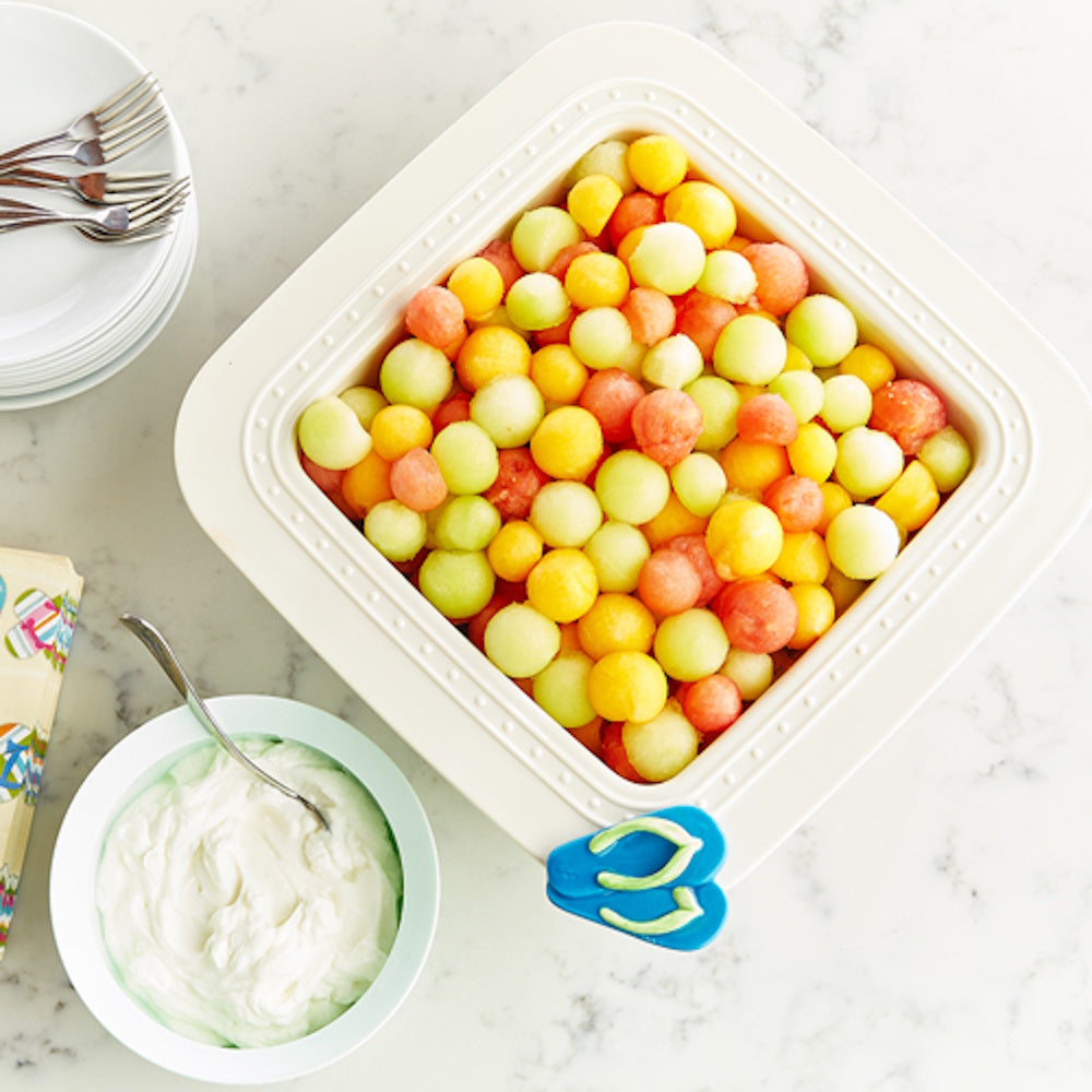 A square white dish filled with colorful melon balls next to a bowl of white dip and a stack of plates.