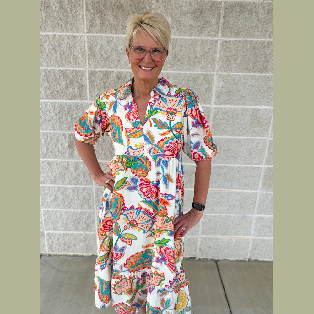A smiling woman with short blonde hair and glasses poses in a colorful, vibrant paisley print midi dress.