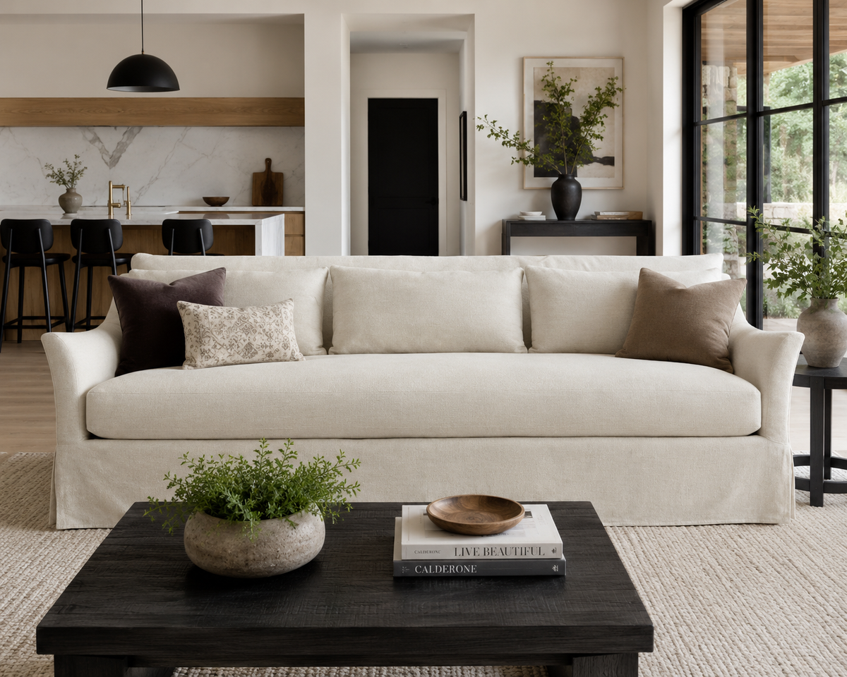 Moreau cream slipcovered sofa in a modern living room with a black coffee table and open kitchen behind it.