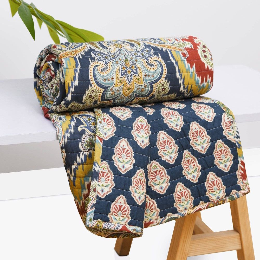Rolled blue quilted throw with colorful paisley and ikat patterns draped over a white table and wooden stool.