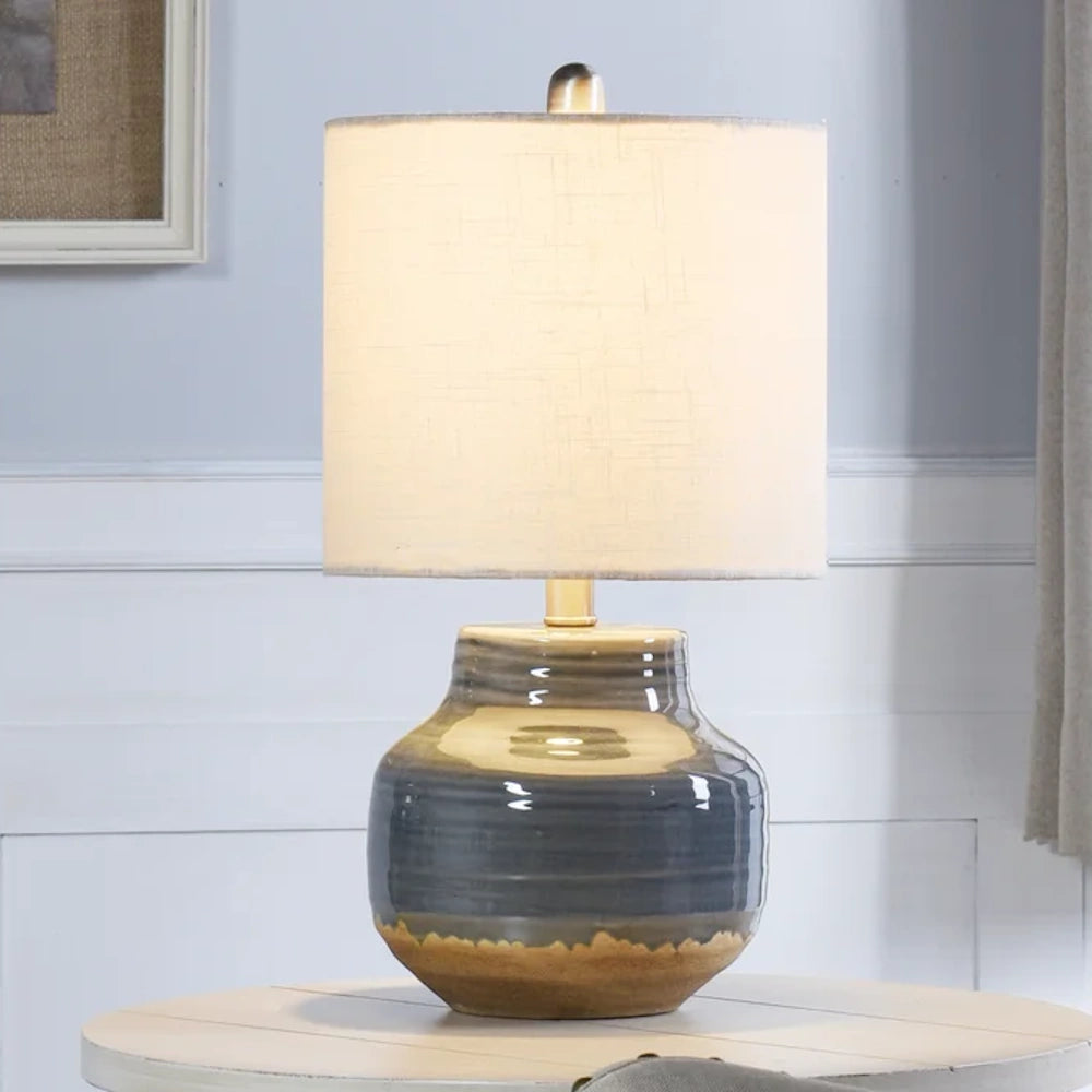 Modern ceramic table lamp with a gray and tan glazed base and a white linen drum shade on a wooden side table.
