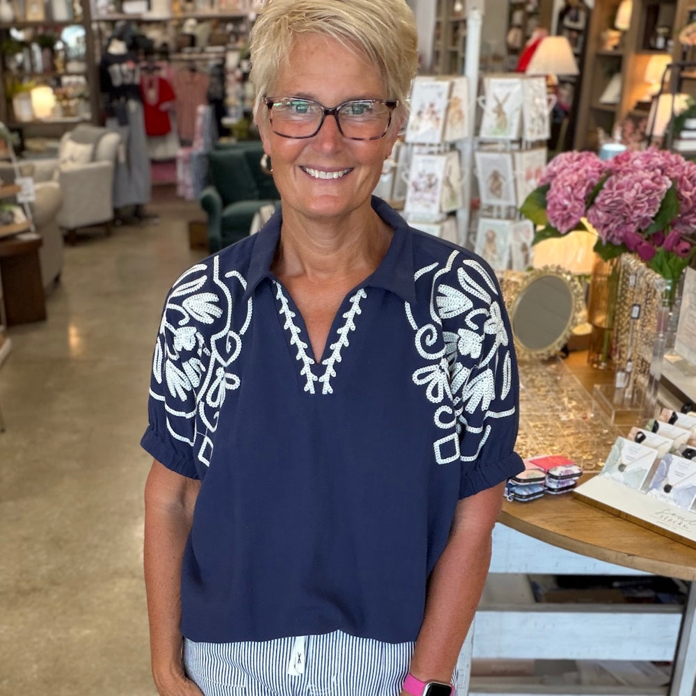 Woman smiles in a navy embroidered v-neck open point collared dolman top with white floral detailing on sleeves.