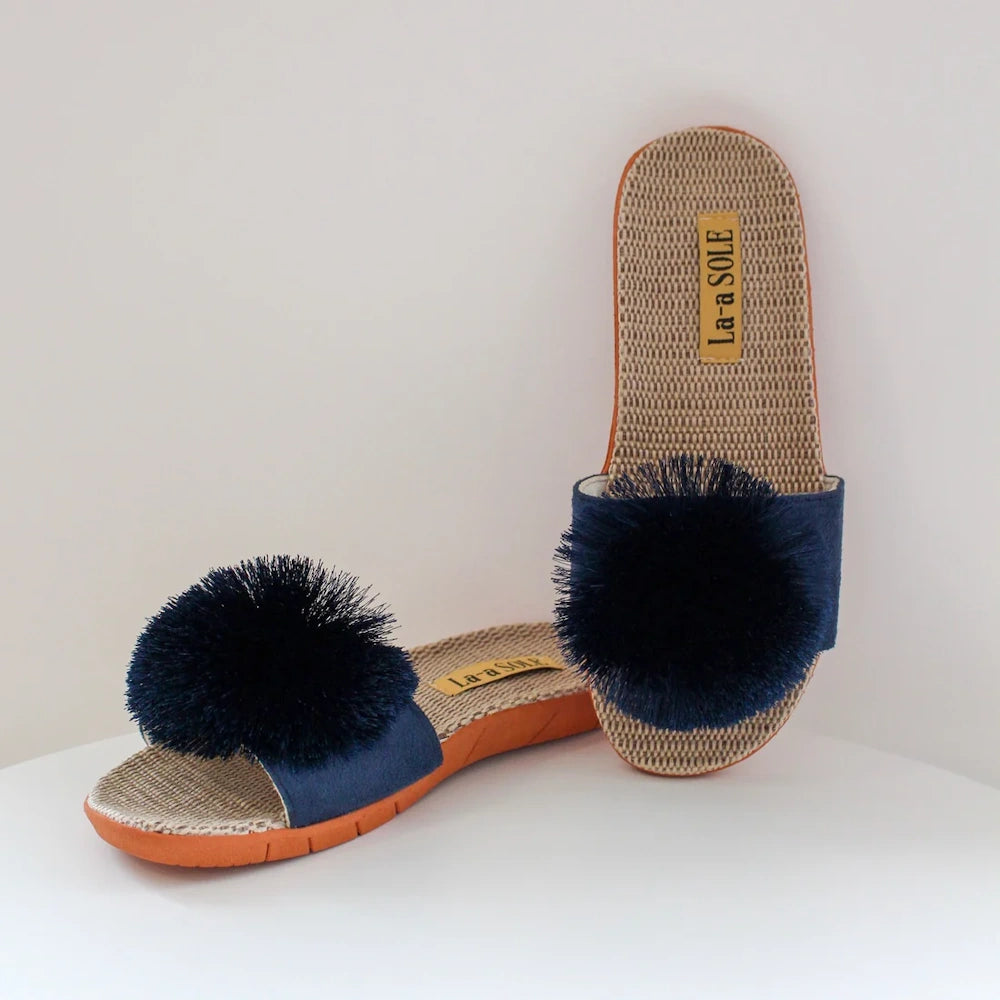 Navy blue slide sandals with large, dark blue pom-poms on the straps and textured tan insoles.