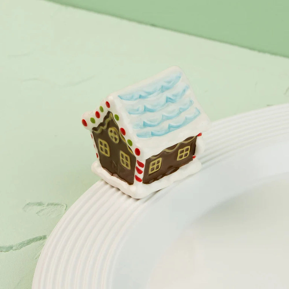 Ceramic gingerbread house figurine with a light blue roof, sitting on the rim of a white textured plate.