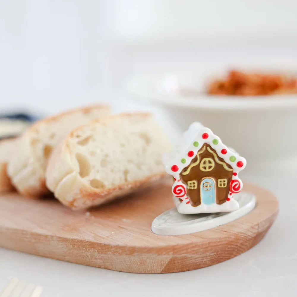 A miniature ceramic gingerbread house figurine attached to a wooden serving board next to sliced bread.