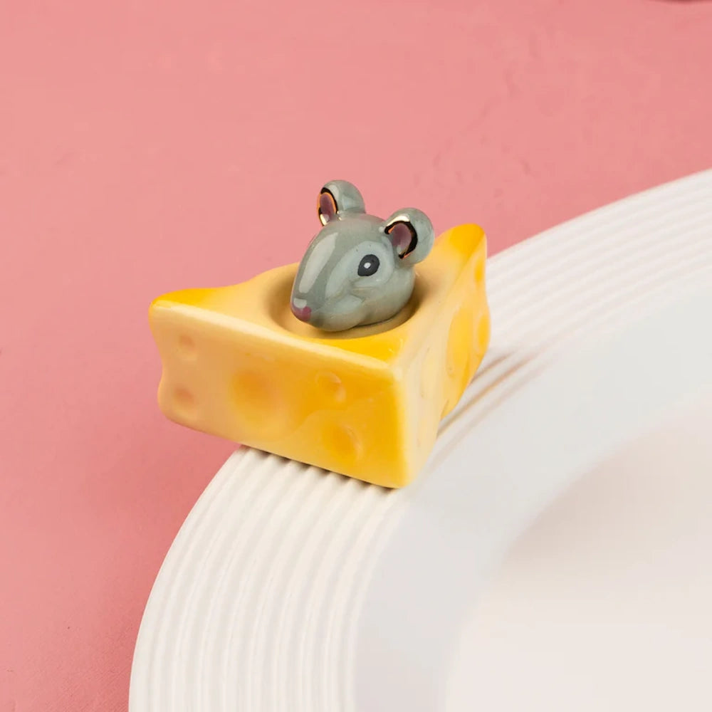 Small ceramic mouse peeking out of a Swiss cheese wedge, attached to the edge of a white plate.