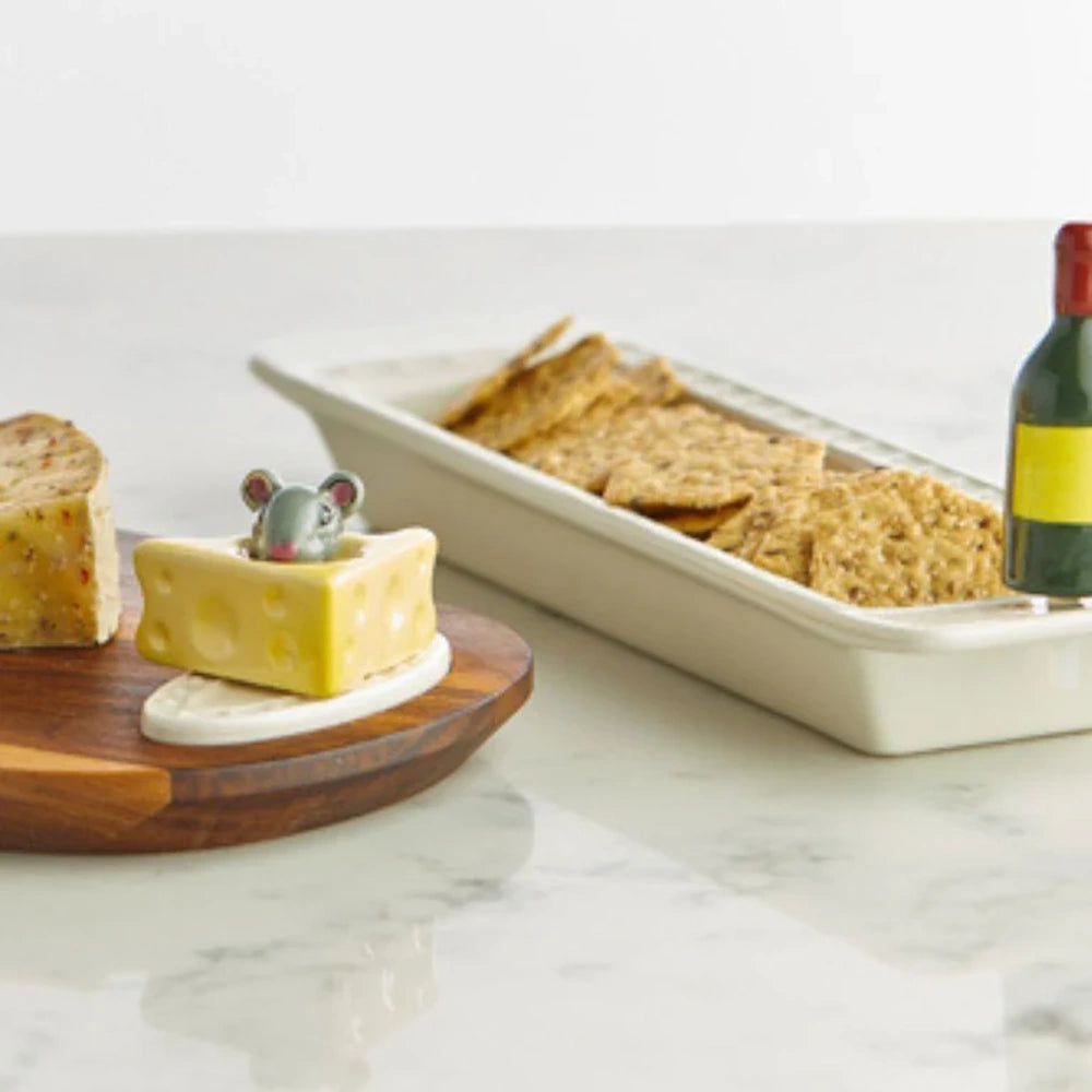 A tiny ceramic mouse popping out of a yellow cheese wedge, served next to crackers, cheese, and a wine bottle.