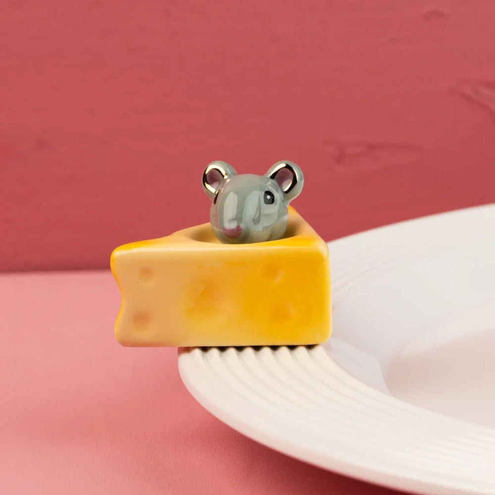 A small ceramic gray mouse peeking out from a yellow wedge of cheese, attached to the rim of a white plate.