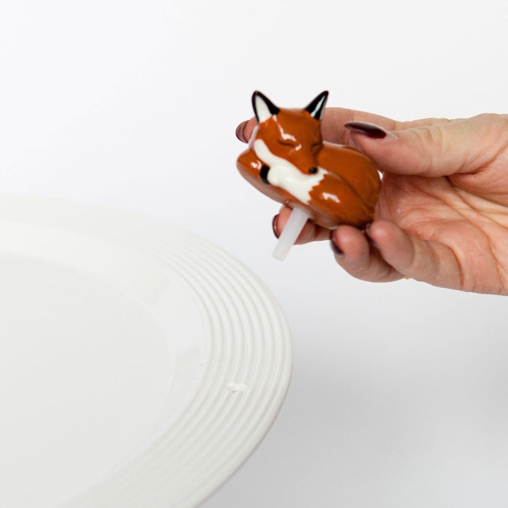 Small ceramic sleeping fox figurine with a peg held by a hand over a white plate rim.
