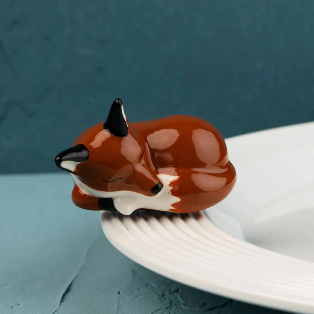 A small ceramic sleeping red fox figurine resting on the rim of a white textured platter, blue background.