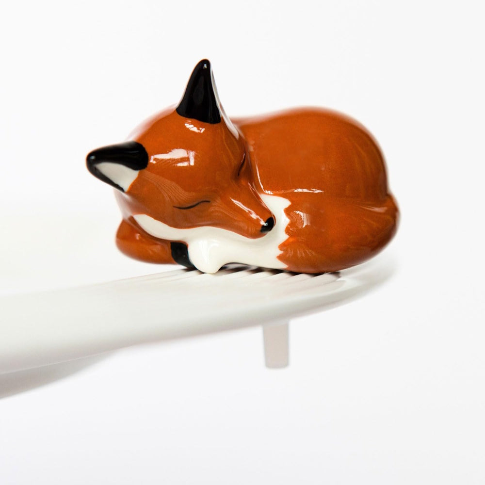 A glossy ceramic figurine of a sleeping orange fox, curled up on the rim of a white ceramic dish.