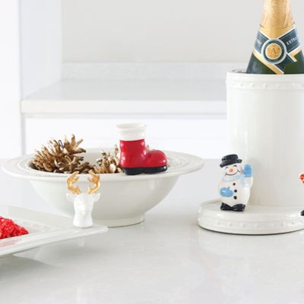 A ceramic snowman figurine stands next to a wine chiller on a white counter decorated with holiday items.