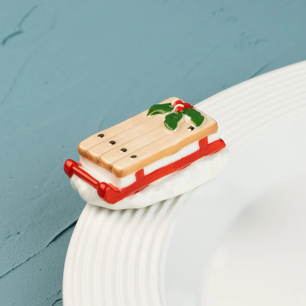 Ceramic wooden sled figurine with red runners and a holly accent, resting on the rim of a white plate.